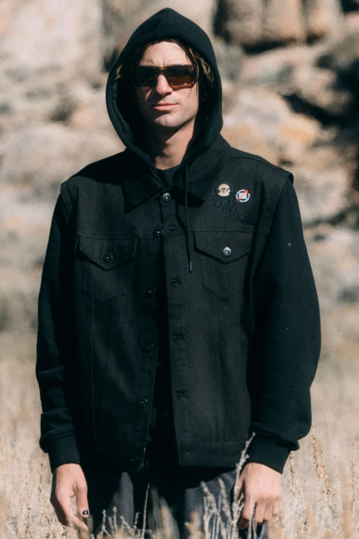 A person wearing a black 686 Men’s Metalhead Trucker Jacket with a built-in hood and two small circular pins on the chest.