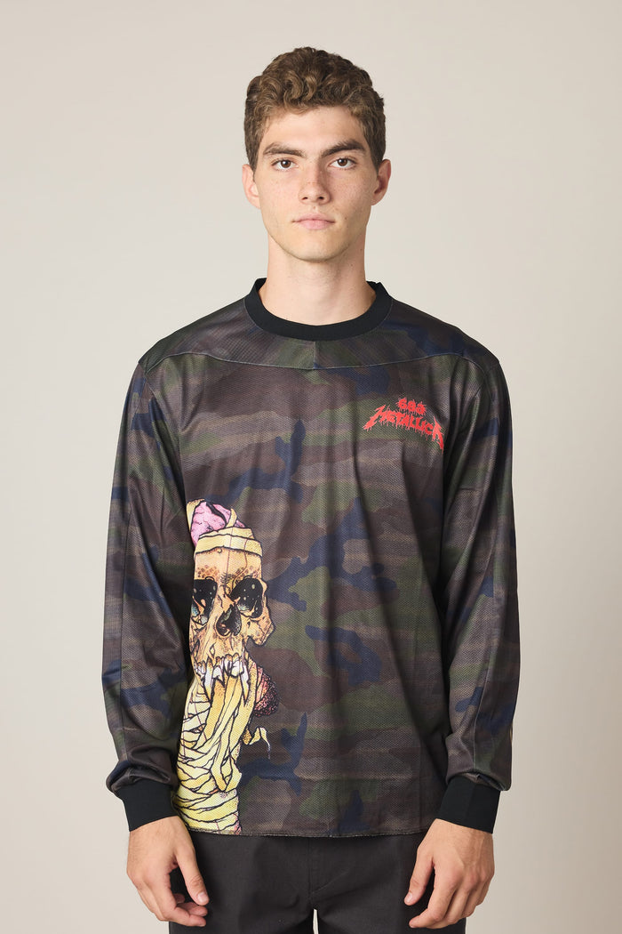 A young man with short curly hair models the 686 Men's Metallica Turbo LS Tech Tee, a long-sleeve, camouflage-style shirt with a bold skull graphic and red Santa Cruz text on the upper left chest, standing against a plain light background.