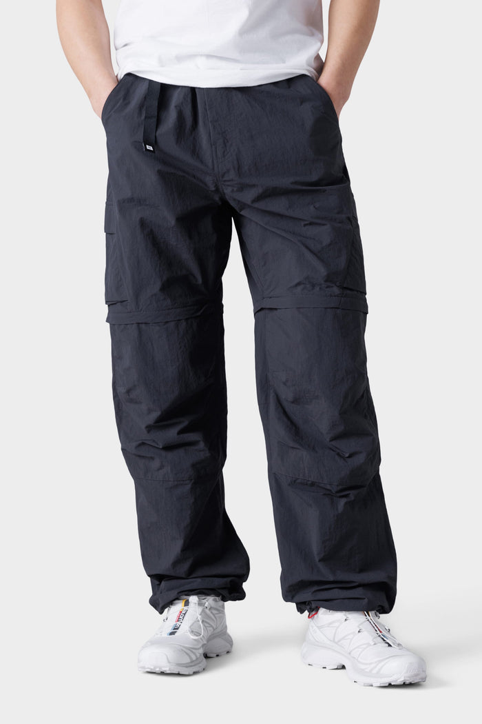 A person wearing 686 Mens Traverse Zip Off Cargo Pant - Wide Fit stands with hands in pockets against a plain light background, paired with a white t-shirt and white sneakers.