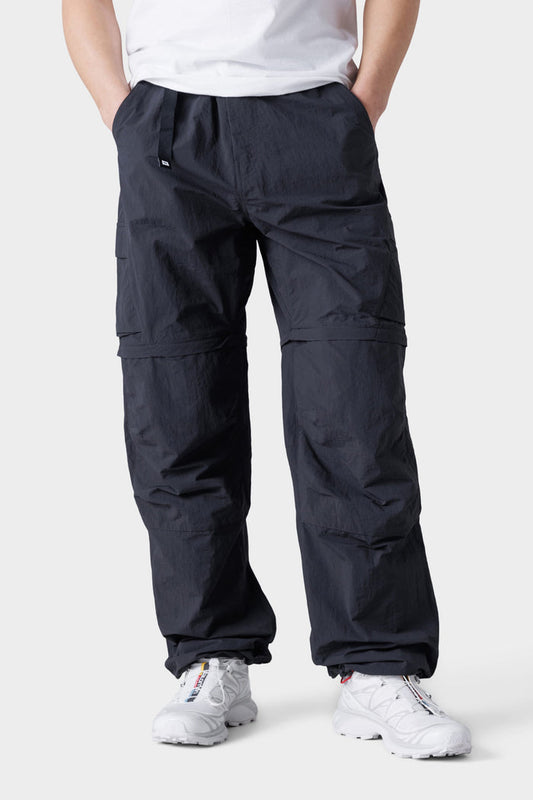 A person wearing 686 Mens Traverse Zip Off Cargo Pant - Wide Fit stands with hands in pockets against a plain light background, paired with a white t-shirt and white sneakers.