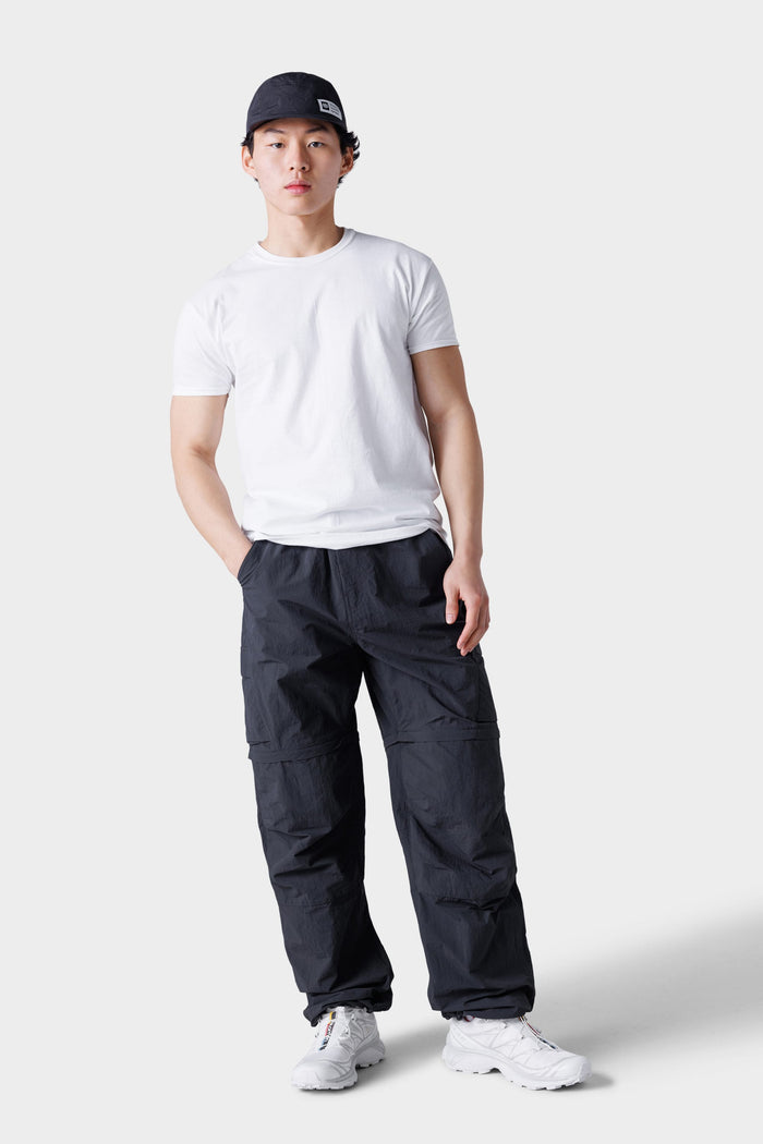 A young man stands against a plain light gray background with one hand in his pocket, wearing a black cap, white t-shirt, white sneakers, and 686 Mens Traverse Zip Off Cargo Pant - Wide Fit by 686.