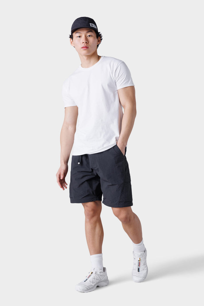 A man wearing 686 Mens Traverse Zip Off Cargo Pant - Wide Fit by 686, a black cap, white T-shirt, white socks, and white sneakers stands against a plain light gray background with one leg slightly bent.