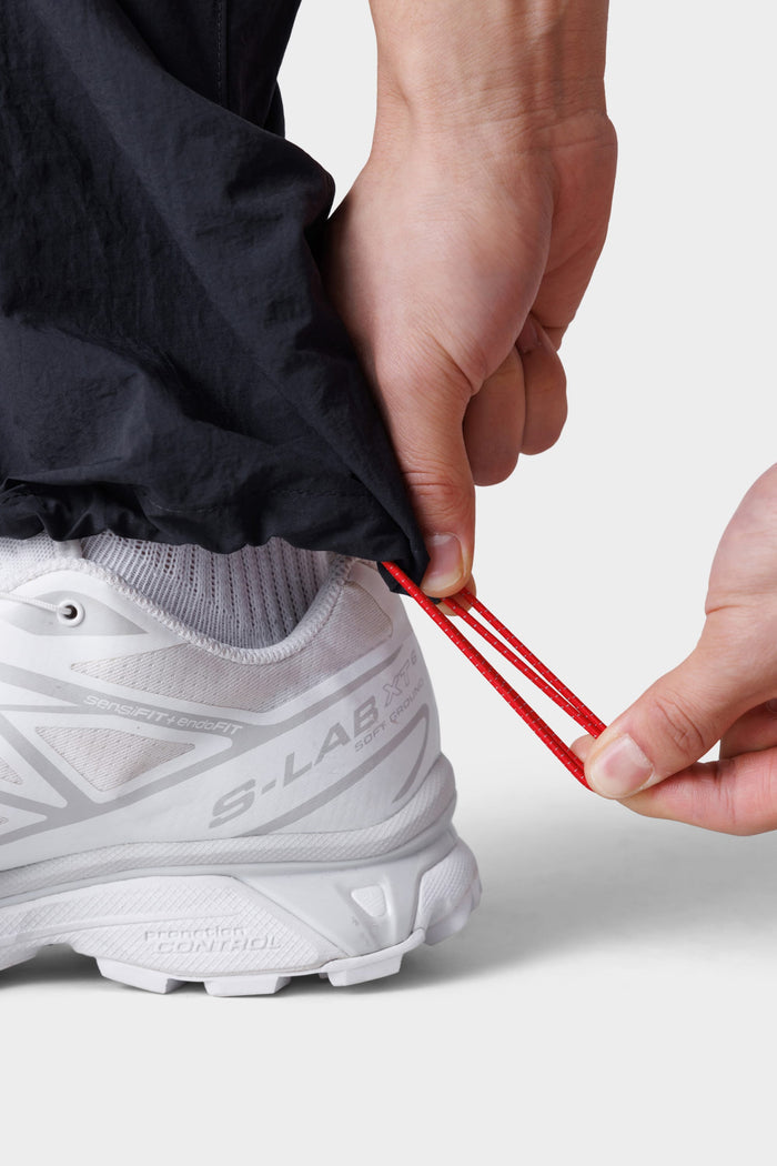 A person adjusts the red ankle drawstring on 686 Mens Traverse Zip Off Cargo Pant - Wide Fit in black, wearing white running shoes, against a plain background.