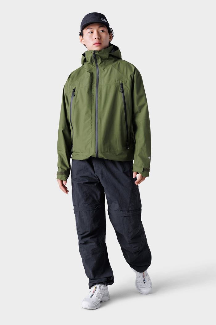 A person in an olive green hooded jacket, white sneakers, a black cap, and 686 Mens Traverse Zip Off Cargo Pant - Wide Fit by 686 stands against a plain light gray background.