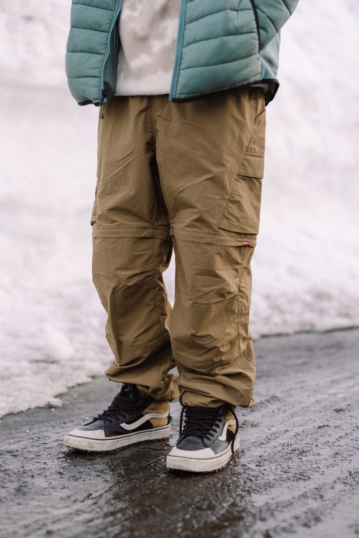 A person stands on a wet, icy path wearing the 686 Mens Traverse Zip Off Cargo Pant - Wide Fit by 686, black and white sneakers, a cream sweater, and a light teal puffer jacket. Snow is piled up in the background.