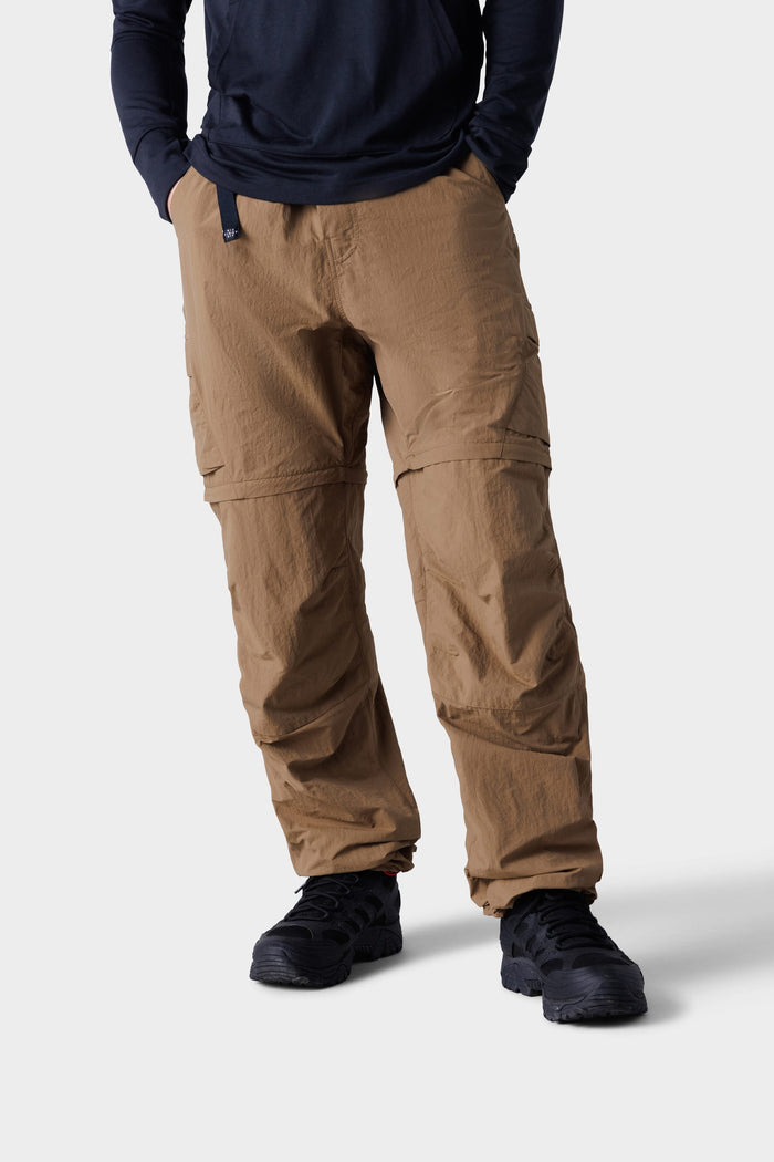 A man in 686 Mens Traverse Zip Off Cargo Pant - Wide Fit (tan), black sneakers, and a navy long-sleeve shirt stands with hands in pockets against a plain light background.