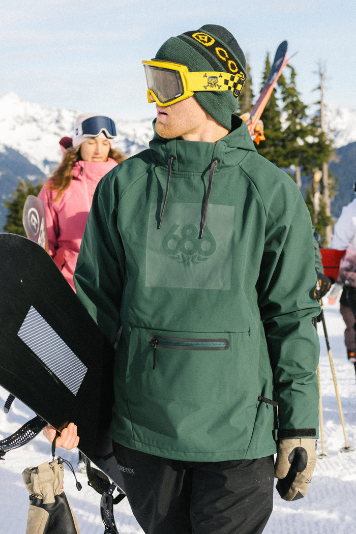 Wearing a 686 Men’s Waterproof Hoody, a person in yellow goggles holds a snowboard on a snowy mountain, with other skiers and snowboarders in the background.