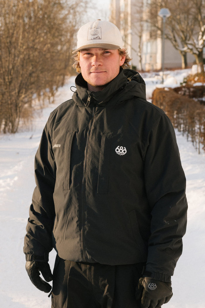 A person wearing the 686 Men’s WINDSTOPPER® Guideline Insulated Jacket by GORE-TEX® LABS stands outdoors on a snowy path, dressed in black gloves and a white cap, with bare trees and buildings visible in the background.