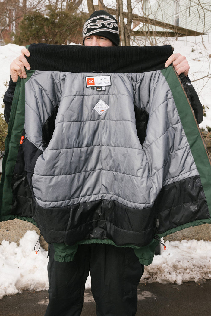 A person holds open the 686 Men’s WINDSTOPPER® Guideline Insulated Jacket by GORE-TEX® LABS outdoors in winter, displaying its quilted interior and labels, with snow and trees in the background.