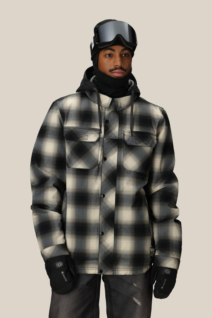 A person stands facing the camera against a plain beige background, wearing the 686 Men’s Woodland™ Insulated Jacket from 686 in a black-and-white plaid pattern, paired with black ski goggles, gloves, a black hood, and neck gaiter.