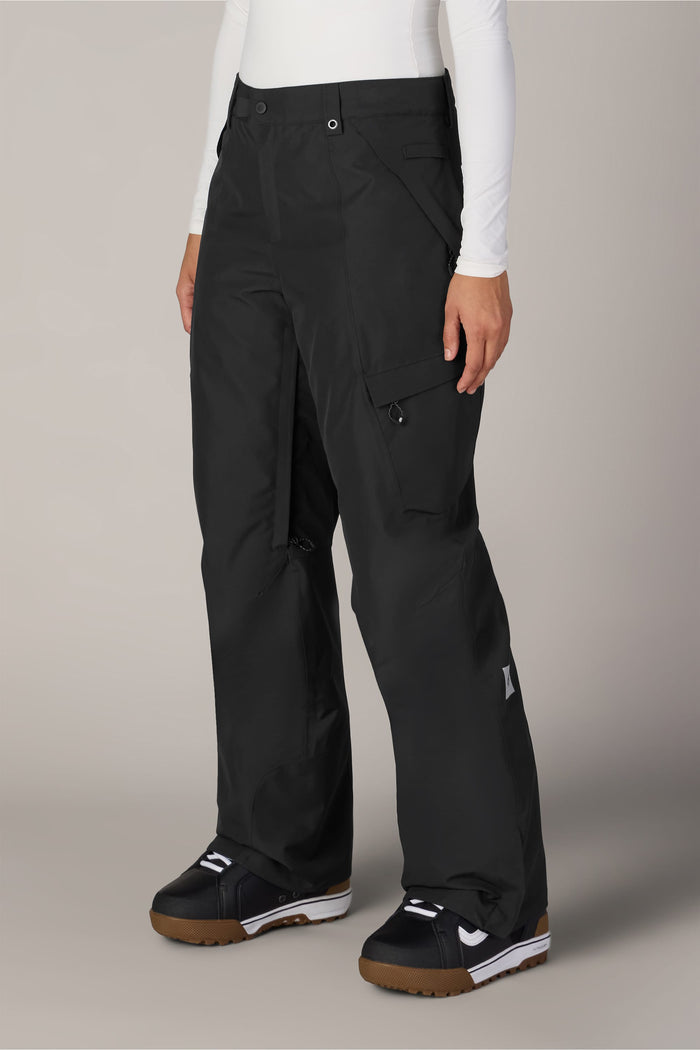 A person is wearing the 686 Women’s Geode Thermagraph® Pant in black, styled as cargo snow pants, paired with a white long-sleeve top and black snow boots with brown soles, standing against a neutral background.