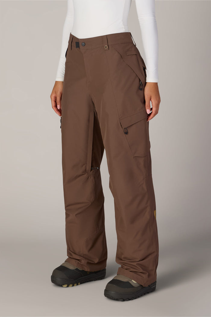 A person wearing the 686 Women’s Geode Thermagraph® Pant in brown, paired with a white long-sleeve top and black winter boots, stands against a plain background. The snow pants from 686 feature multiple pockets and zippers.