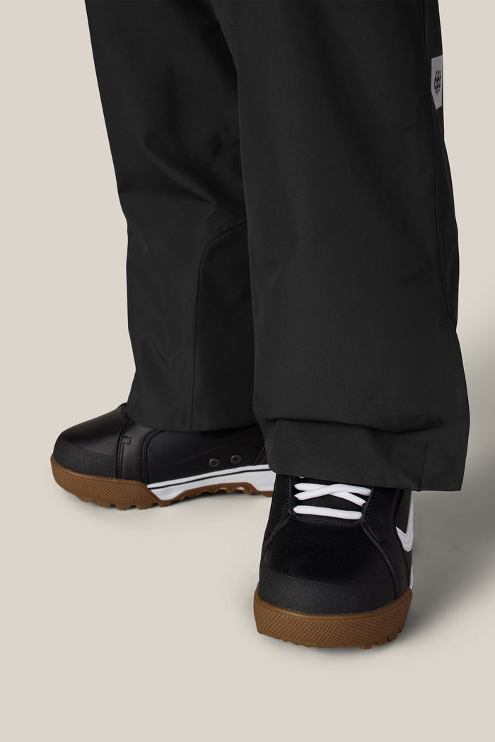 A close-up of a person wearing the 686 Women’s Geode Thermagraph® Pant, paired with black sneakers featuring white laces and brown soles, standing on a light beige background.