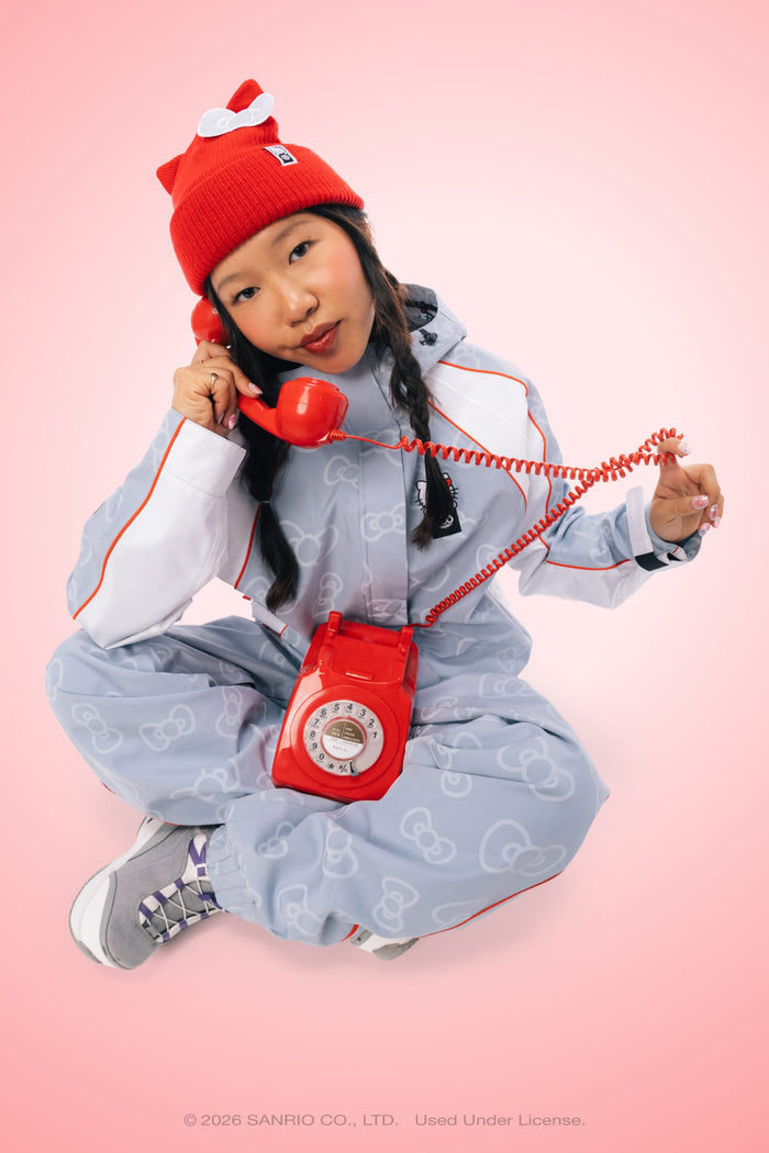 A person in a light blue Hello Kitty jumpsuit and the 686 Women’s Hello Kitty® Knit Beanie by 686 sits cross-legged, holding a red rotary phone against a pink background.