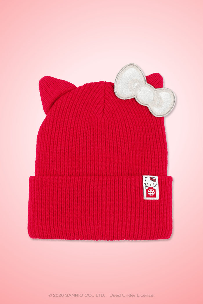 The 686 Women’s Hello Kitty® Knit Beanie by 686 features a red knit design with cat ears, a white bow on one ear, and a small Hello Kitty tag on the brim, displayed against a pink background.