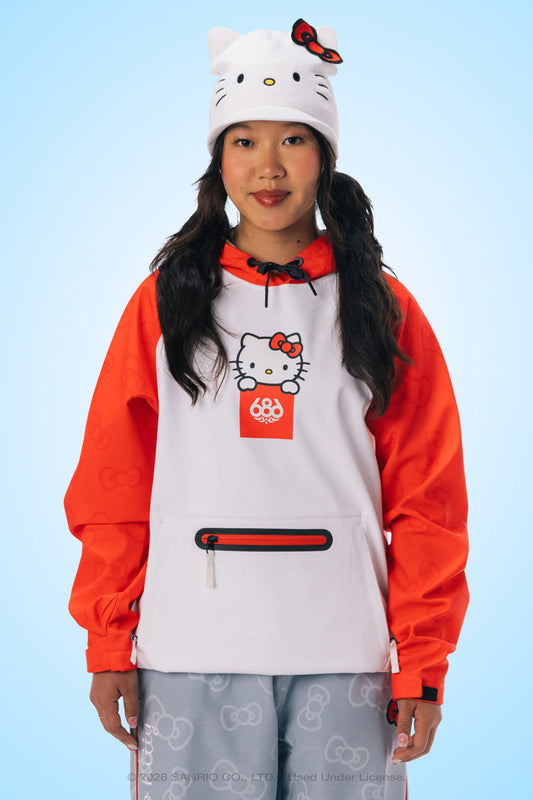 A person is wearing a white Hello Kitty hat, the 686 Women’s Hello Kitty® Waterproof Hoody by 686—a white and red woven hoodie with a zipper pocket—and light blue pants with subtle Hello Kitty patterns, standing against a light blue background.