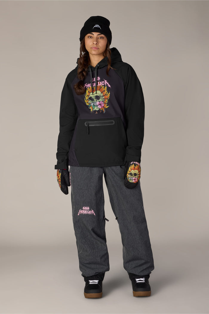 A person wearing a 686 Women’s Metallica Waterproof Hoody in black polyester and matching dark gray snow pants, both featuring Metallica graphics, with a black beanie and gloves.