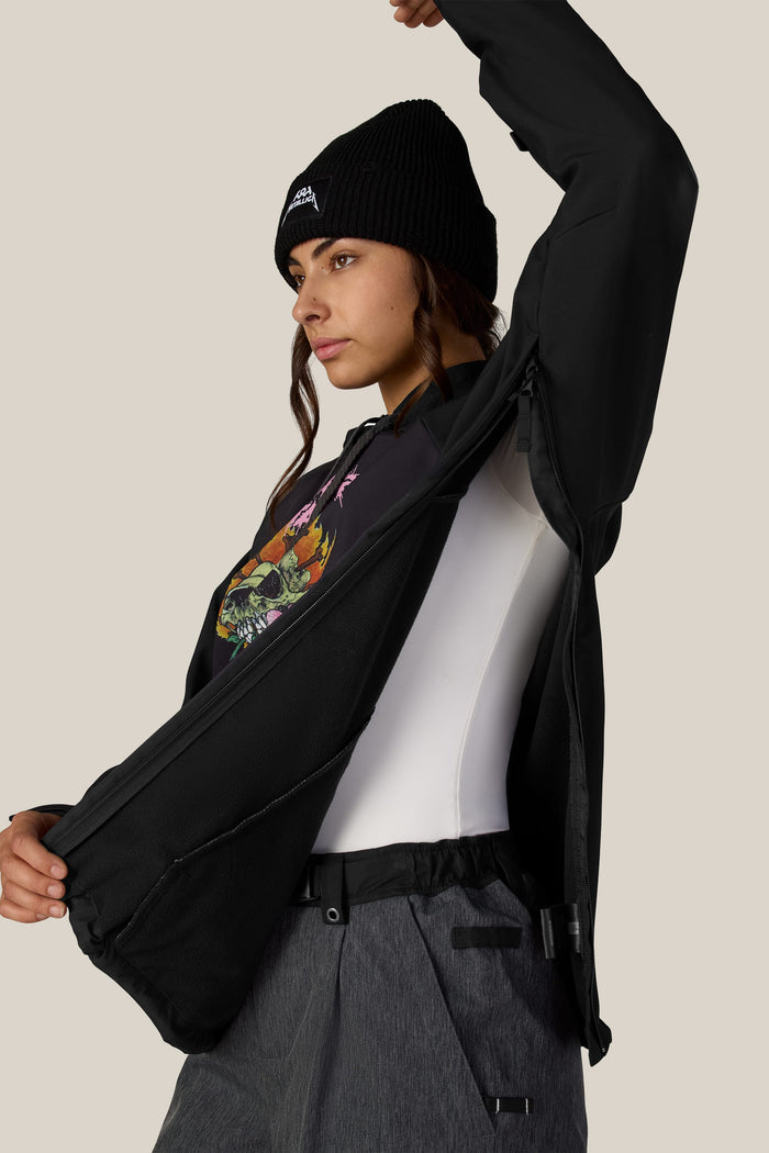 A woman wears a black knit beanie and a 686 Women’s Metallica Waterproof Hoody with side zippers partially opened to reveal a white base layer.