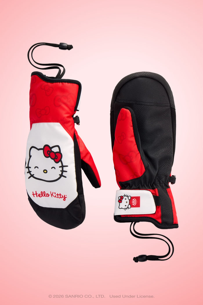 Two 686 Women’s Revel Mitts in red, black, and white—featuring Hello Kitty’s face, a red bow, and “Hello Kitty” text—are displayed on a pink gradient background. Each mitten includes a black wrist strap.