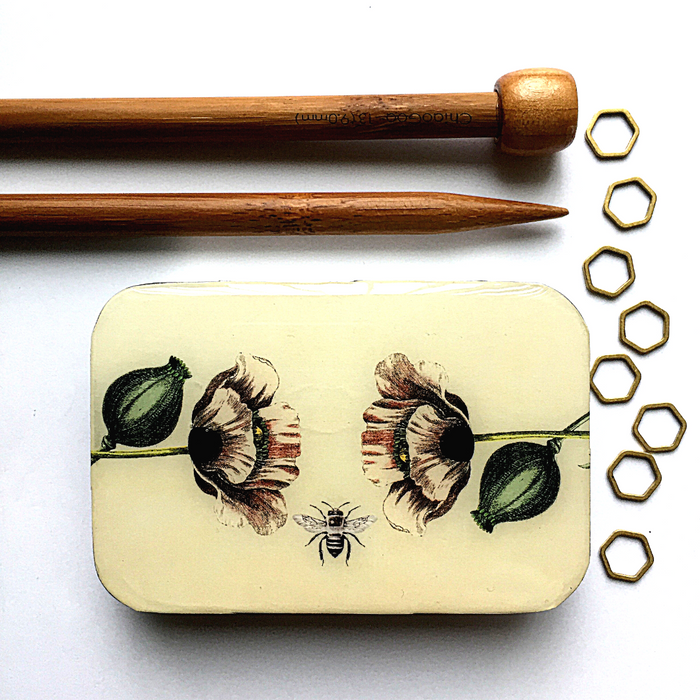 Decorative tin with floral design, wooden knitting needles, and metal hexagonal shapes on a white background
