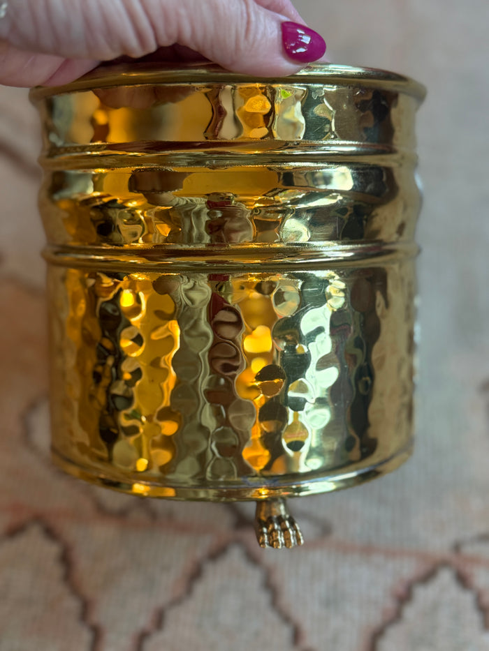 Gold textured container held by a hand with pink nail polish on a patterned background